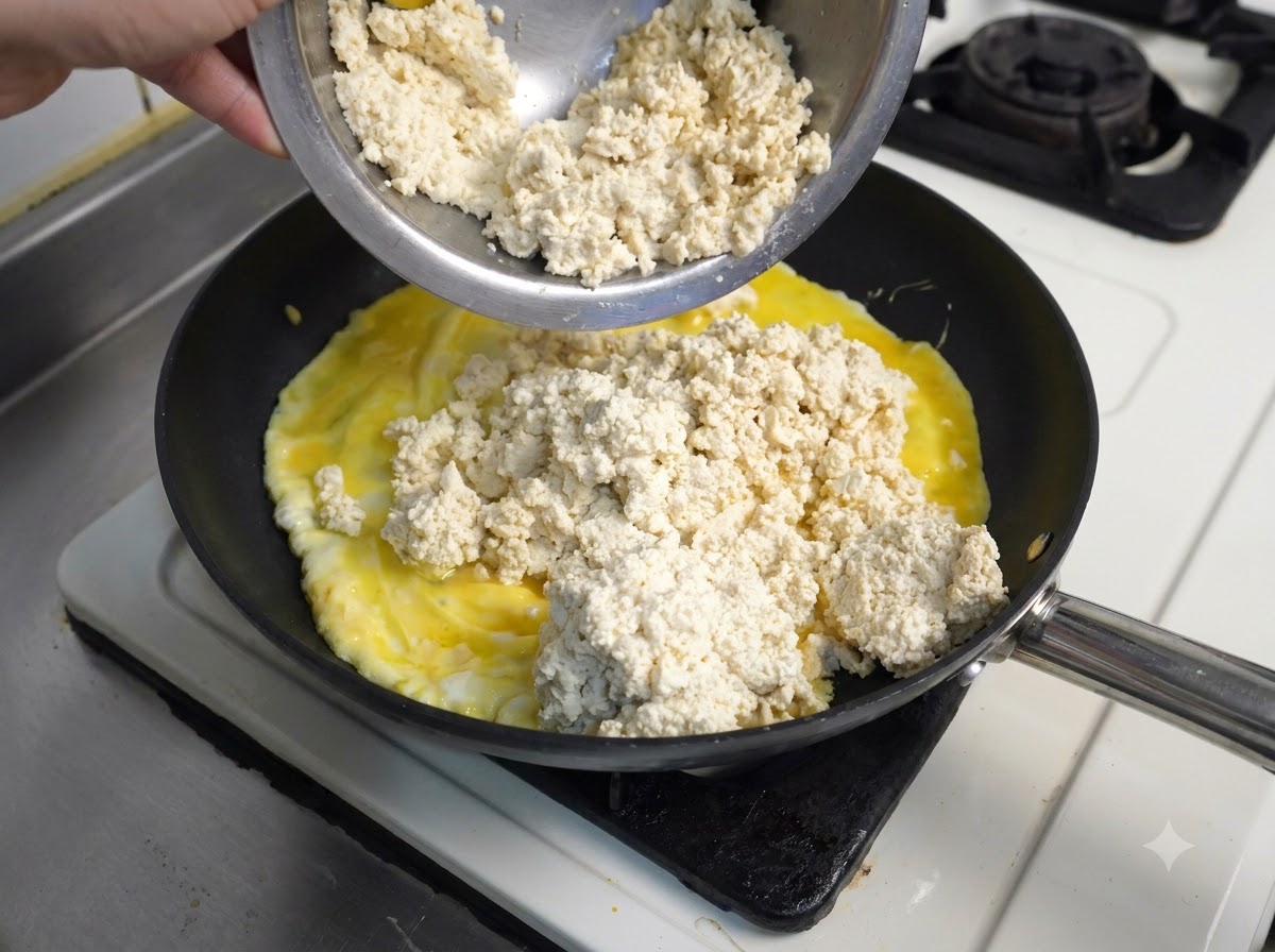 A metal bowl pouring crumbled white tofu over yellow scrambled eggs cooking in a black frying pan.