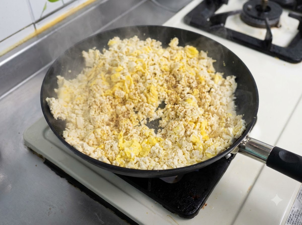 A frying pan filled with a mixture of scrambled eggs and tofu being stir-fried with seasoning sprinkled on top.