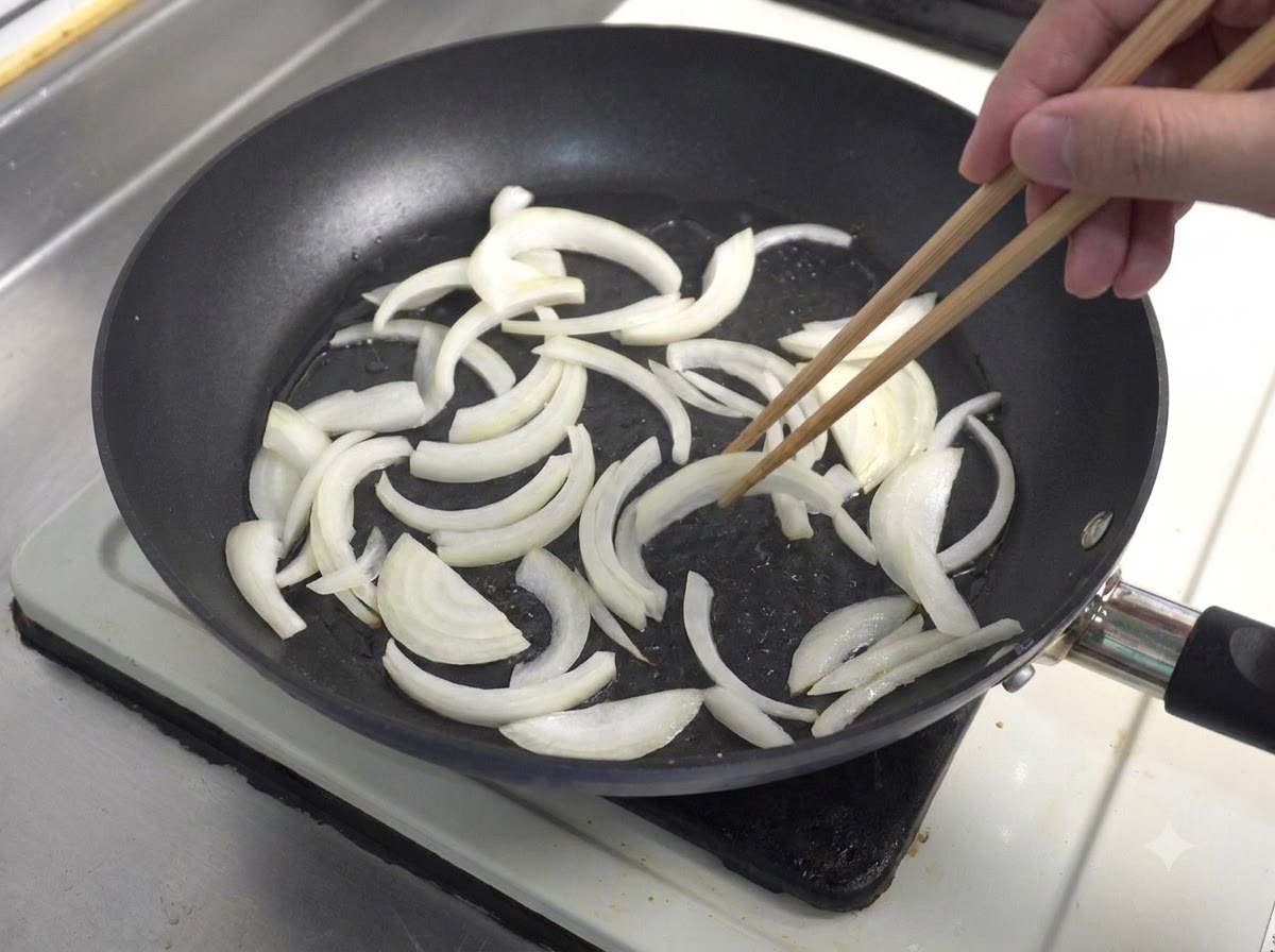 Sliced white onions being sautéed in a frying pan using wooden chopsticks.