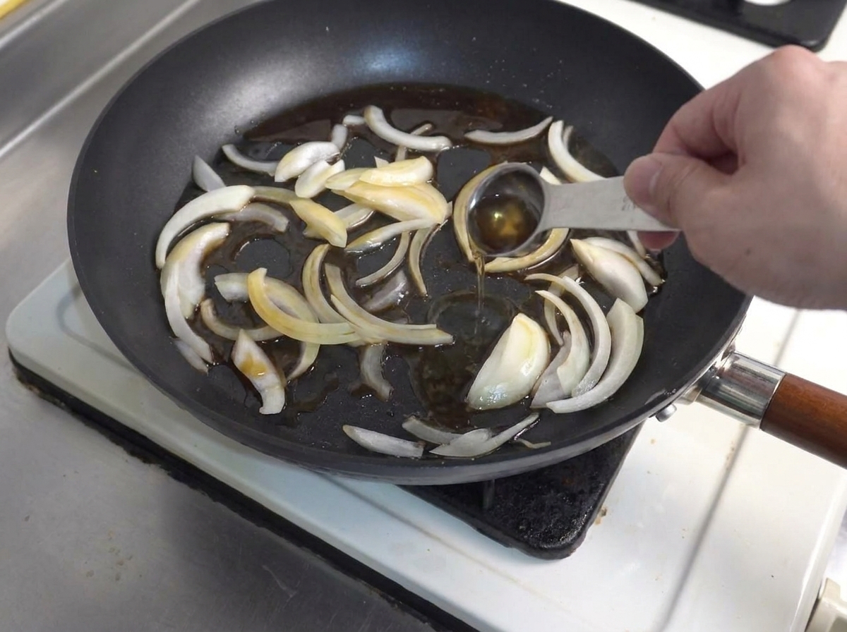 A measuring spoon pouring liquid seasoning into a frying pan with sautéed onion slices.