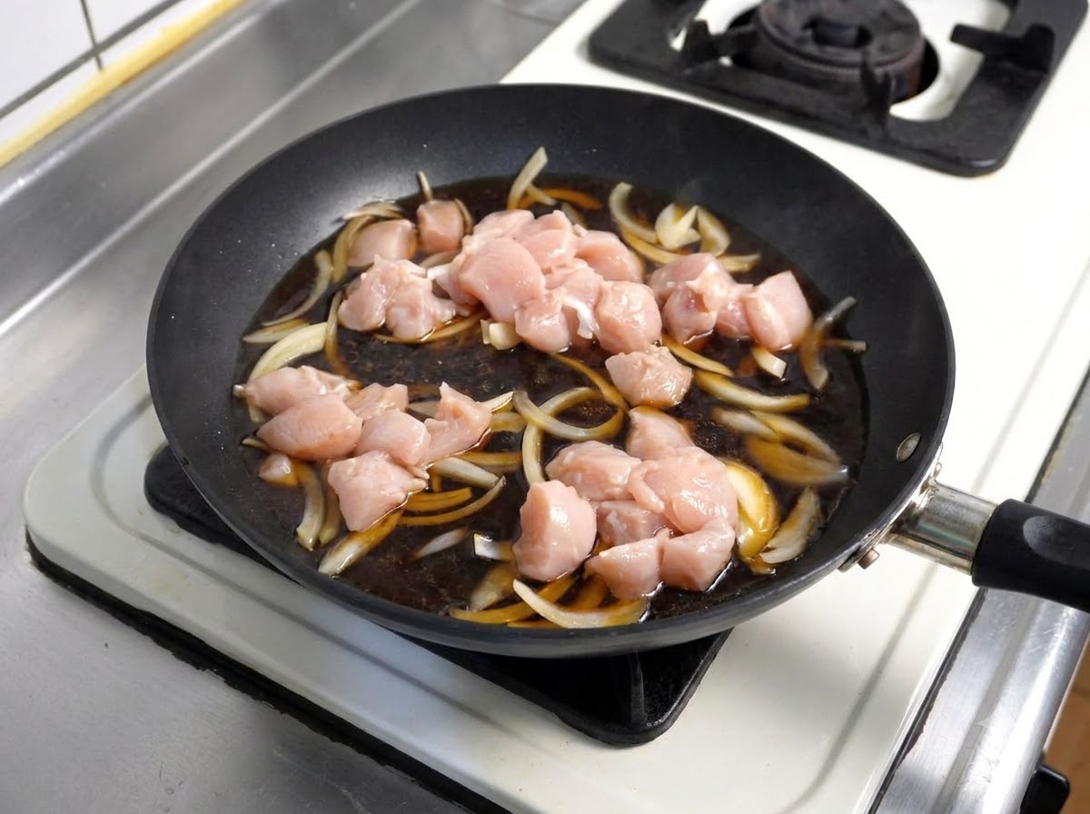 Raw chicken pieces placed into a simmering dark sauce with onions inside a frying pan.