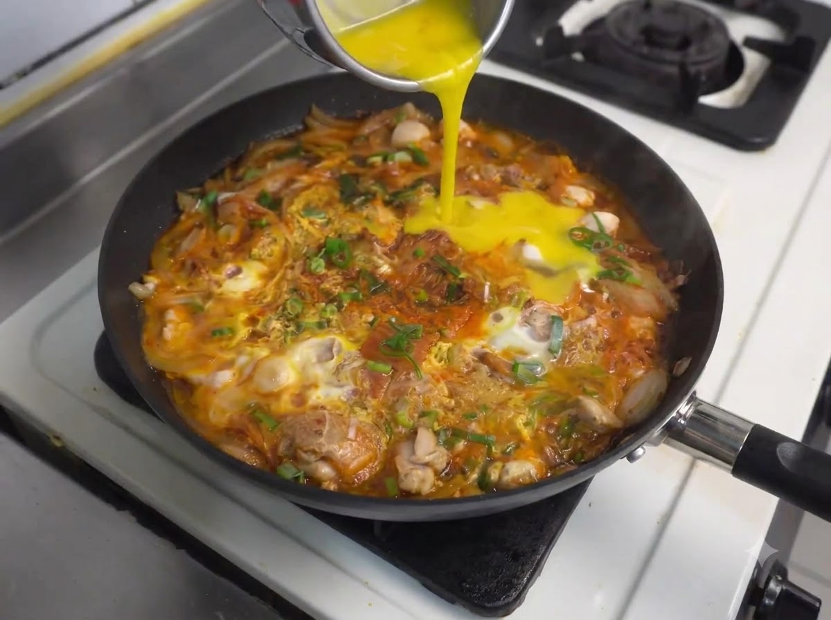 Pouring more beaten eggs from a container into the simmering pan with chicken and kimchi.