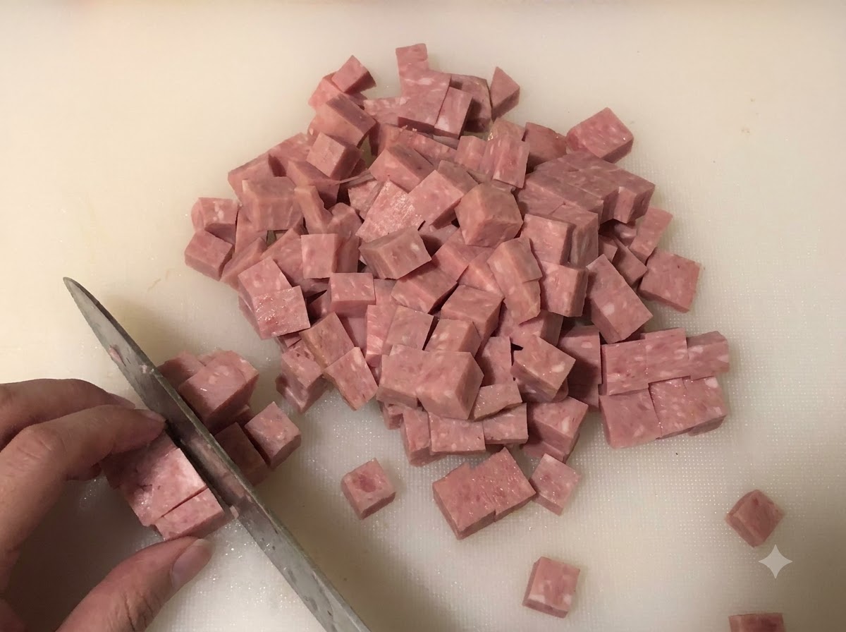 Spam being sliced into small cubes on a cutting board with chopped onions and green onions in the background.