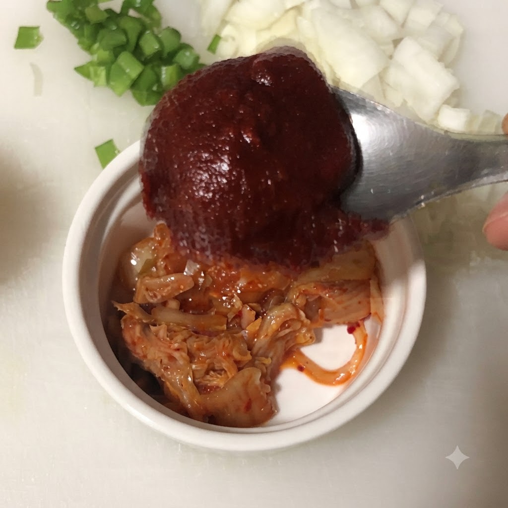 A spoonful of Korean chili paste being held over a bowl containing chopped kimchi.