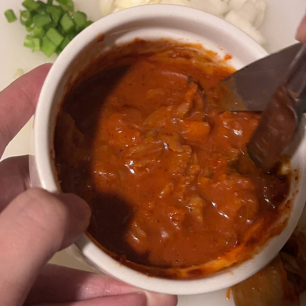Kitchen scissors cutting through kimchi pieces submerged in a thick red sauce mixture in a small white bowl.