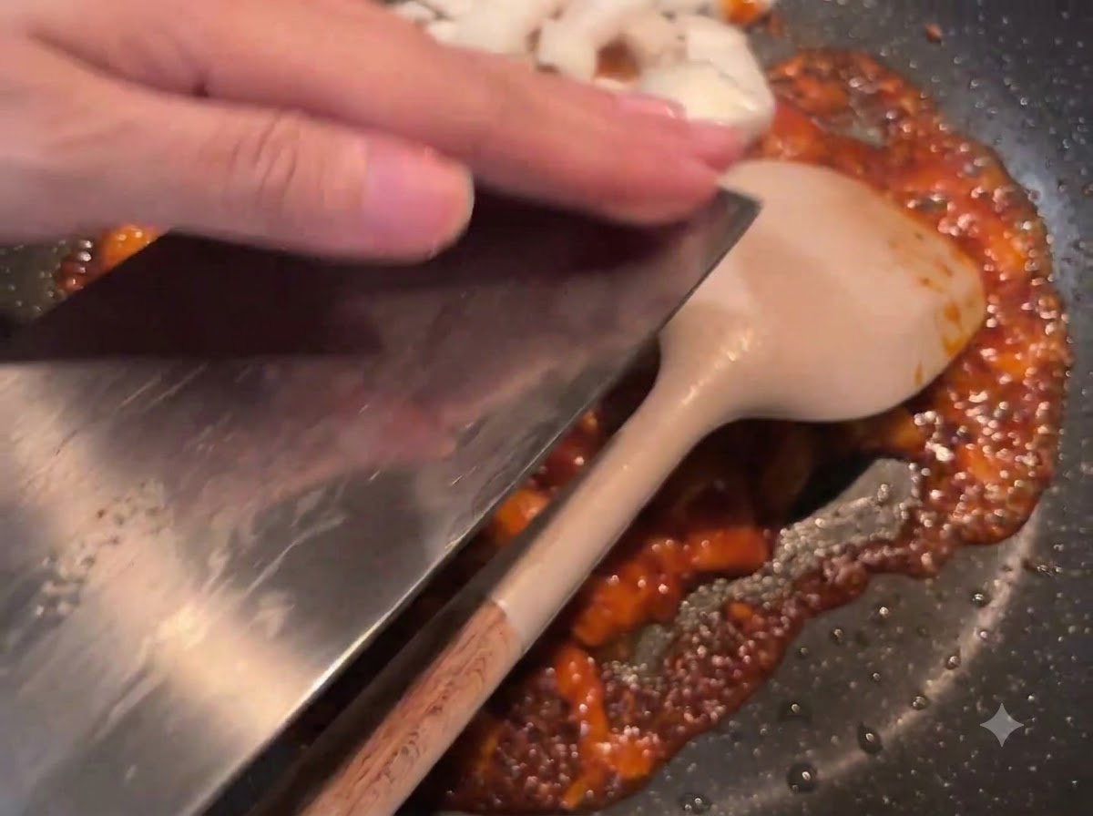 Diced white onions being scraped off a cleaver into the frying pan containing the sautéed red sauce.