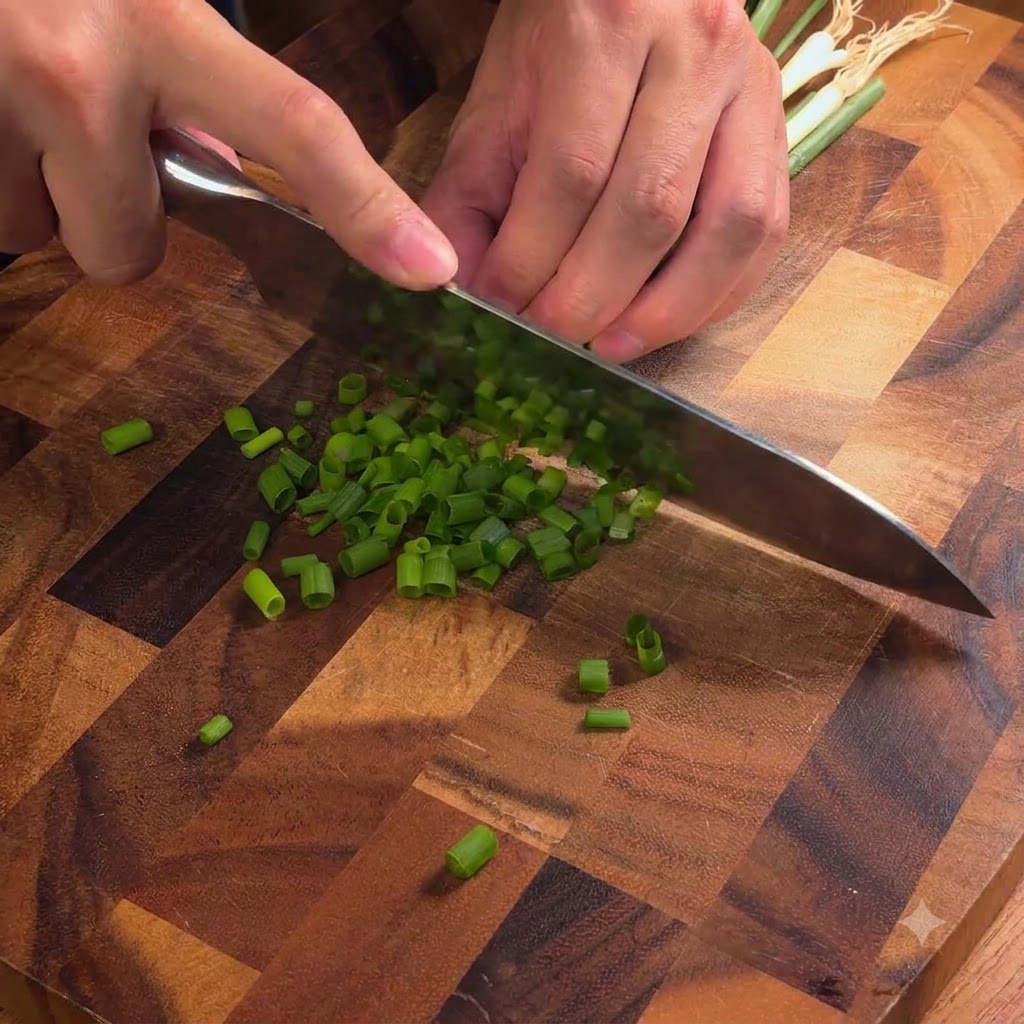 A chefs knife slicing fresh green scallions into fine pieces on a wooden cutting board.