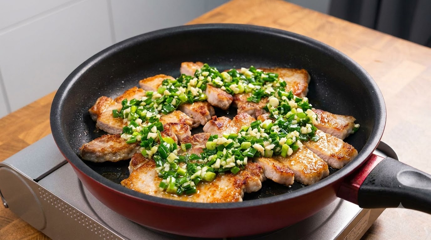 Golden-brown pan-fried pork chop slices generously topped with fresh green scallions and minced garlic in a dark non-stick pan
