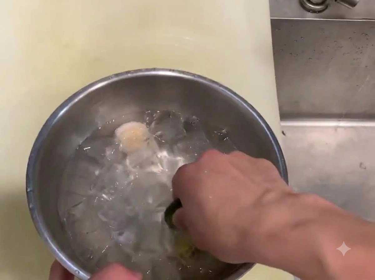 A hand swirling a raw scallop muscle inside a metal bowl filled with ice cubes and cold water.