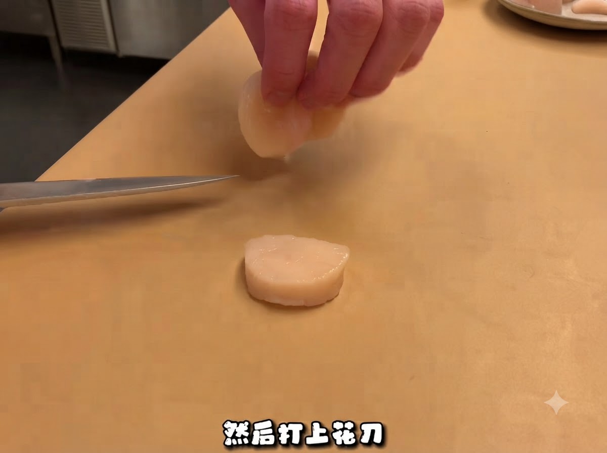 A chefs knife resting near a sliced raw scallop on a cutting board, with hands handling a piece above.