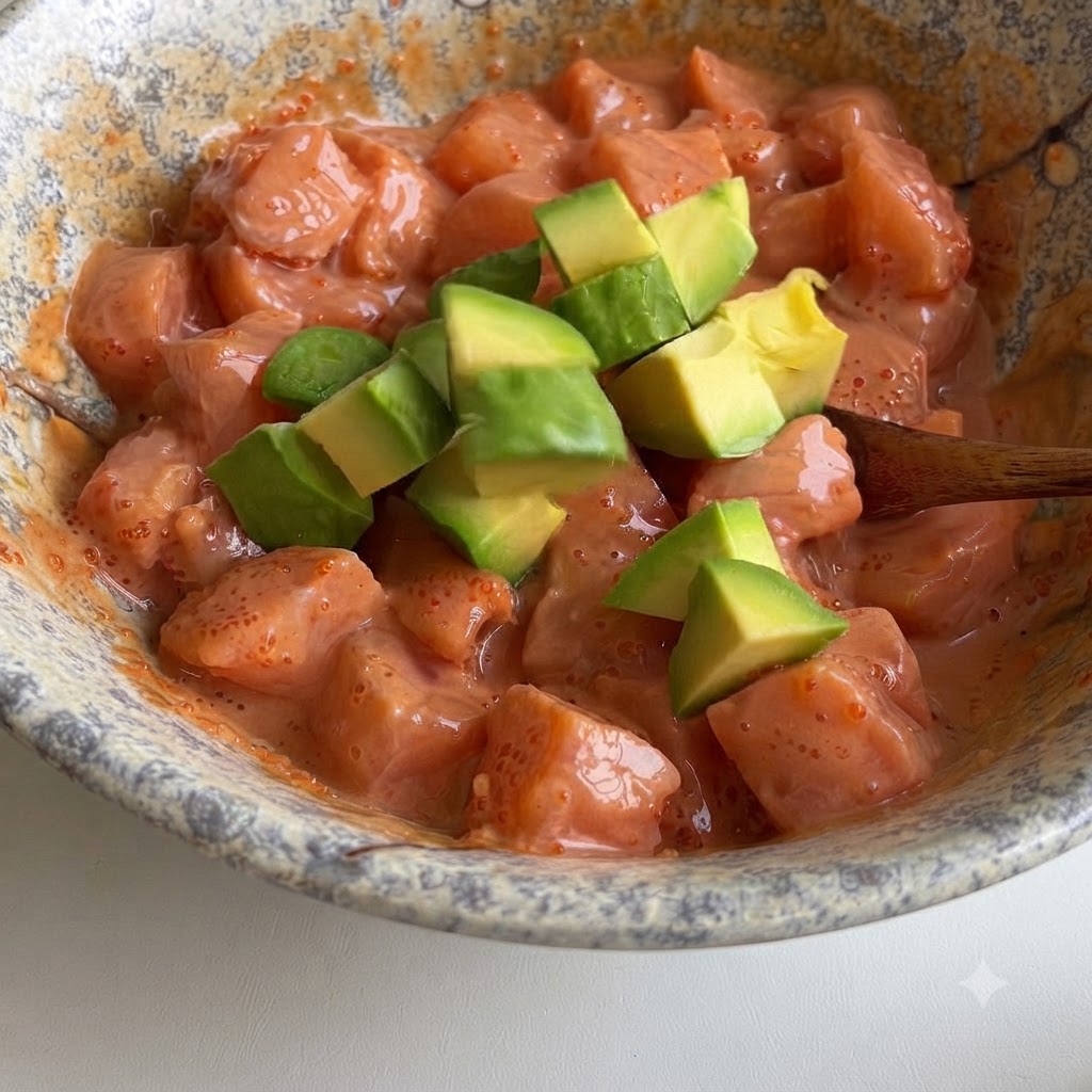 Green avocado cubes being gently folded into a bowl of salmon cubes coated in a creamy pink sauce.