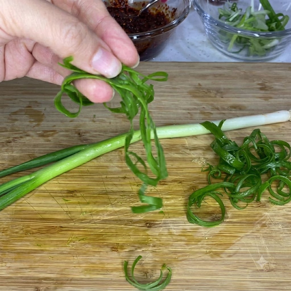A hand holding curled green scallion strips above a wooden cutting board, with glass bowls of sauce and sliced onions in the background.