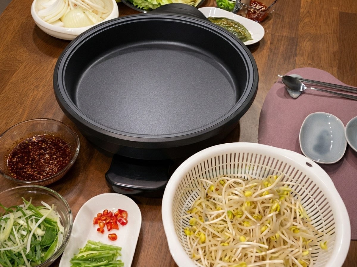 An electric skillet placed on a wooden table alongside a colander of bean sprouts, a bowl of sauce, and chopped vegetables.
