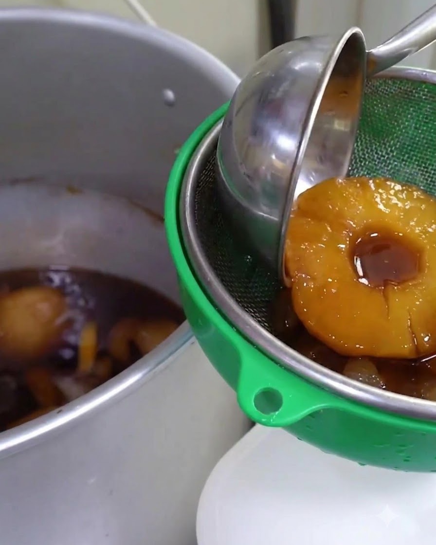 Simmered fruit-infused sauce being strained through a green sieve into a large metal container.