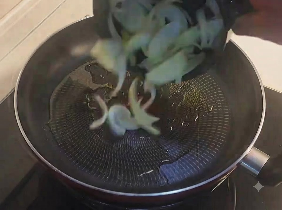 Sliced yellow onions being tossed into an oiled black frying pan.