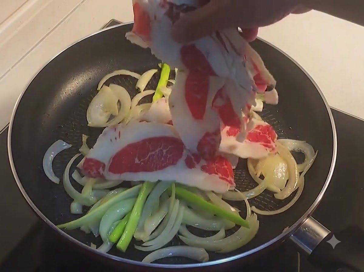 A hand placing thin slices of raw beef belly into a frying pan with sliced onions and green onions.