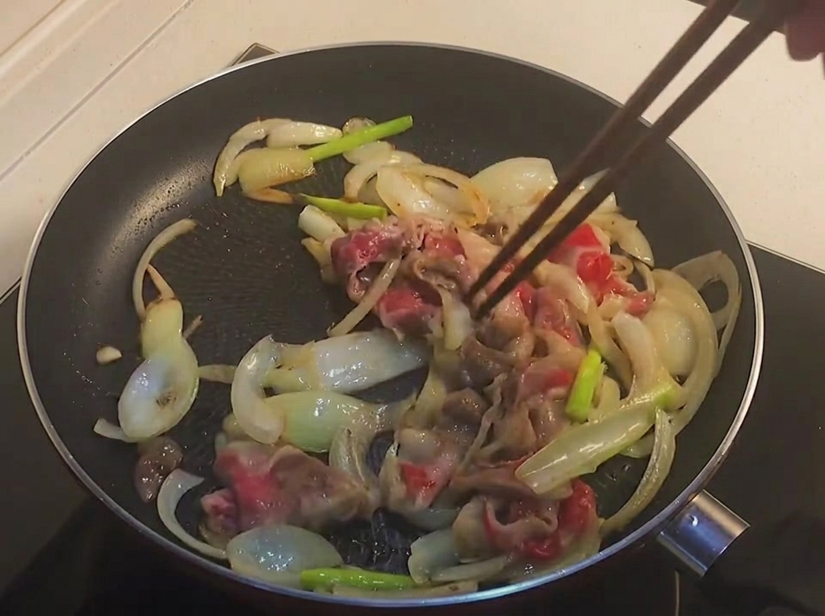 Chopsticks stirring partially cooked, lightly browned beef belly slices and onions in a black frying pan.