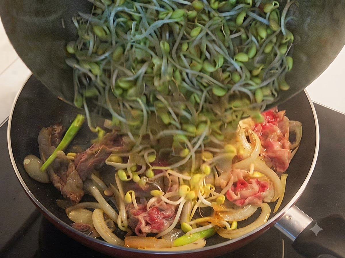 Fresh yellow soybean sprouts being poured from a bowl into a frying pan containing partially cooked beef and onions.