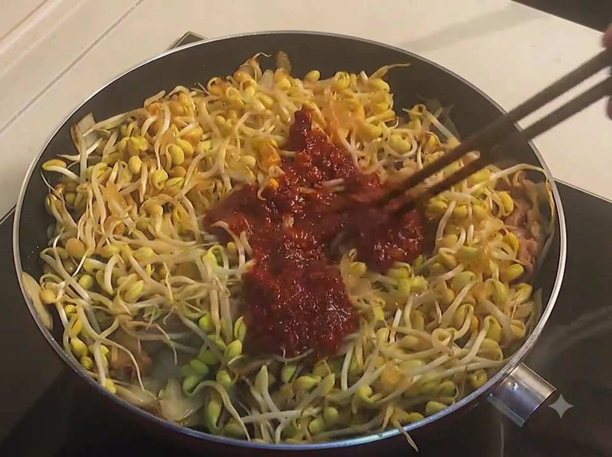 Chopsticks tossing yellow soybean sprouts and beef with a thick, dark red spicy sauce in a pan.