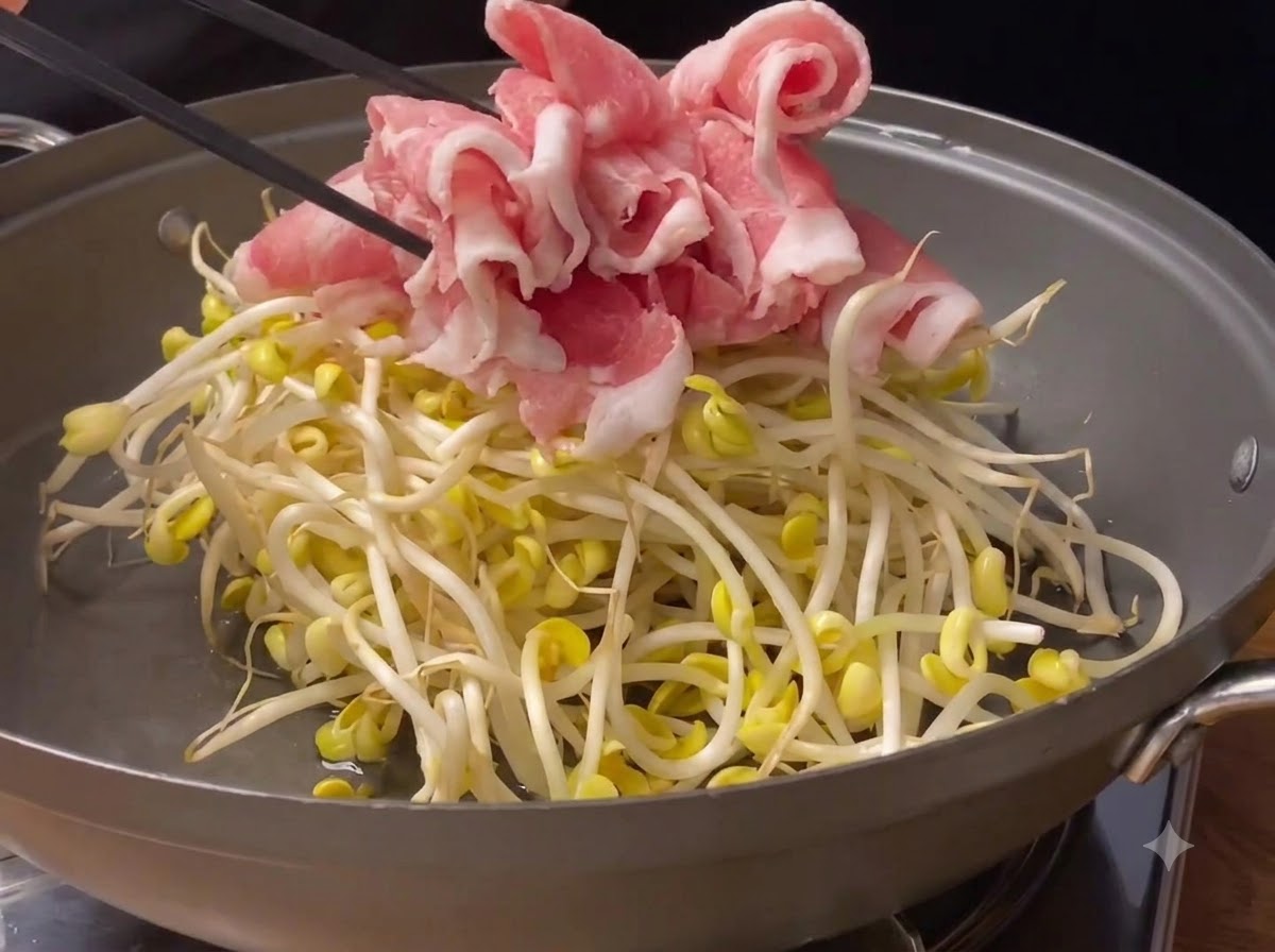 Thinly sliced pink pork rolls being placed on top of fresh soybean sprouts inside a cooking pan.