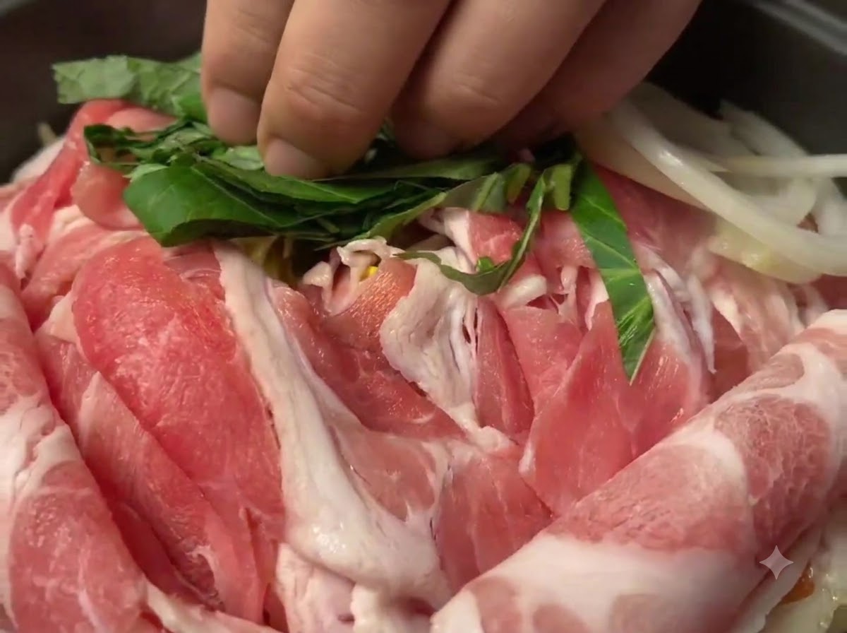 Chopped green perilla leaves being scattered over raw pork slices in the pan.
