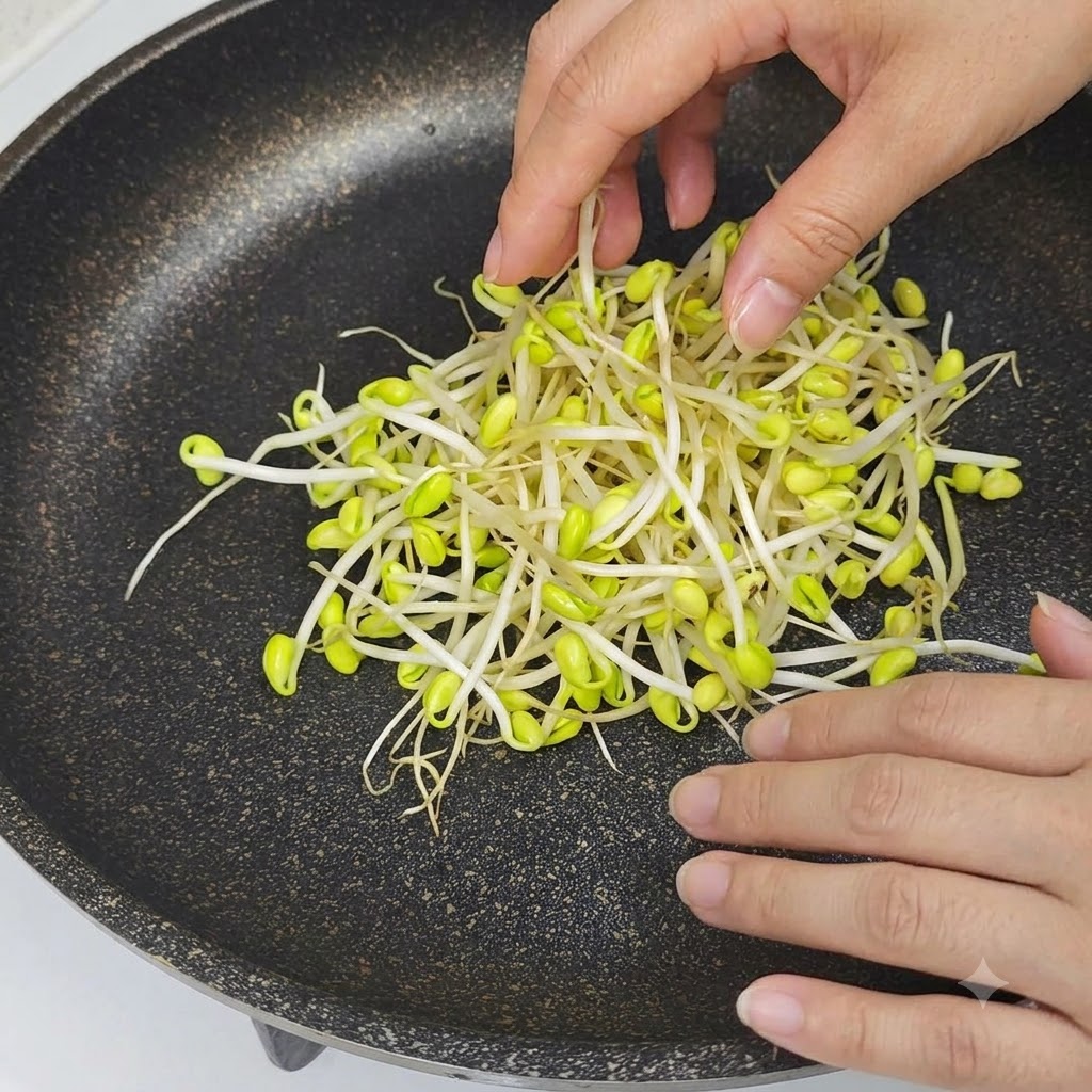 Hands placing raw yellow-tipped bean sprouts into the center of a black non-stick pan.