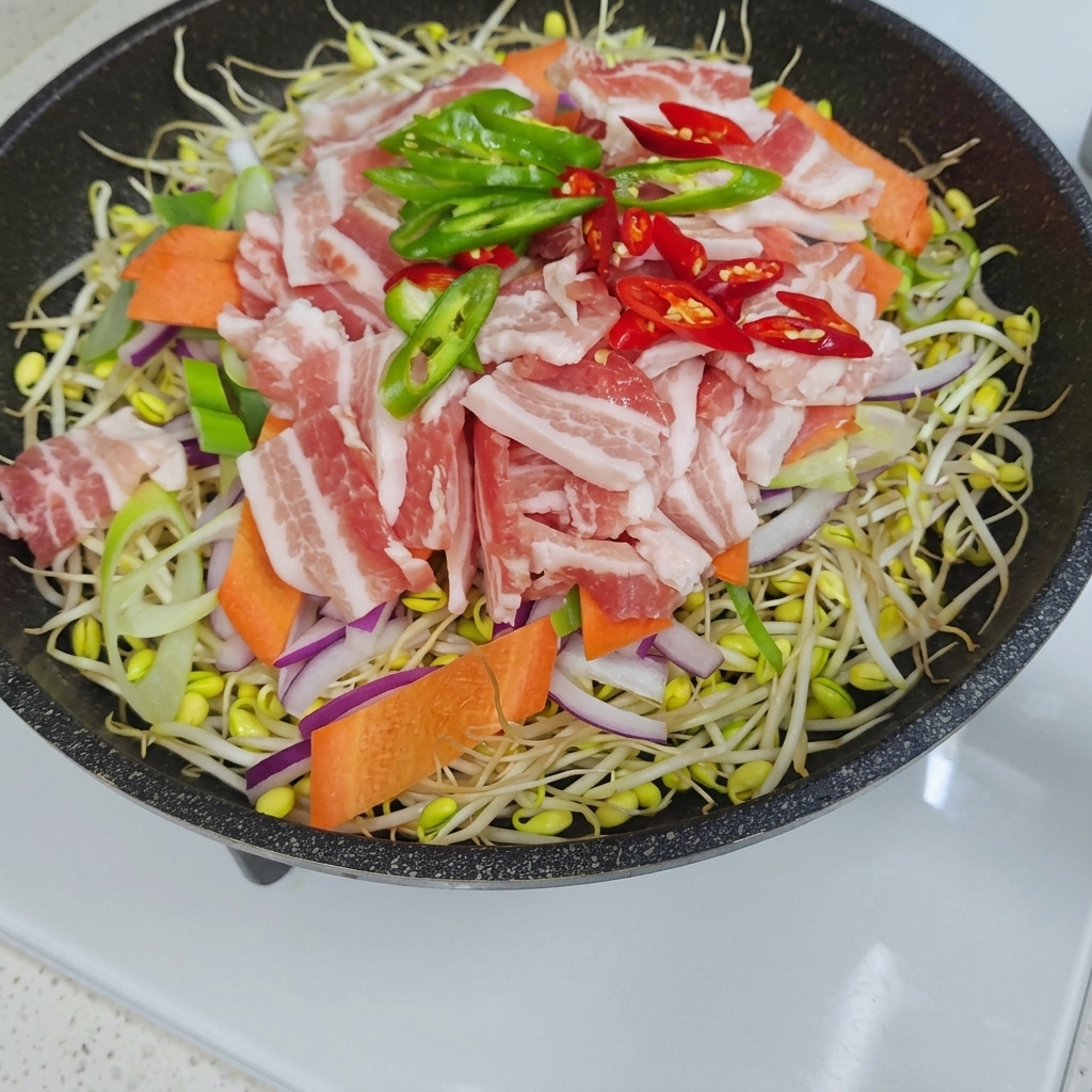 An overhead view of a pan filled with a layer of bean sprouts topped with sliced onions, carrots, raw pork belly strips, and green and red chili slices.