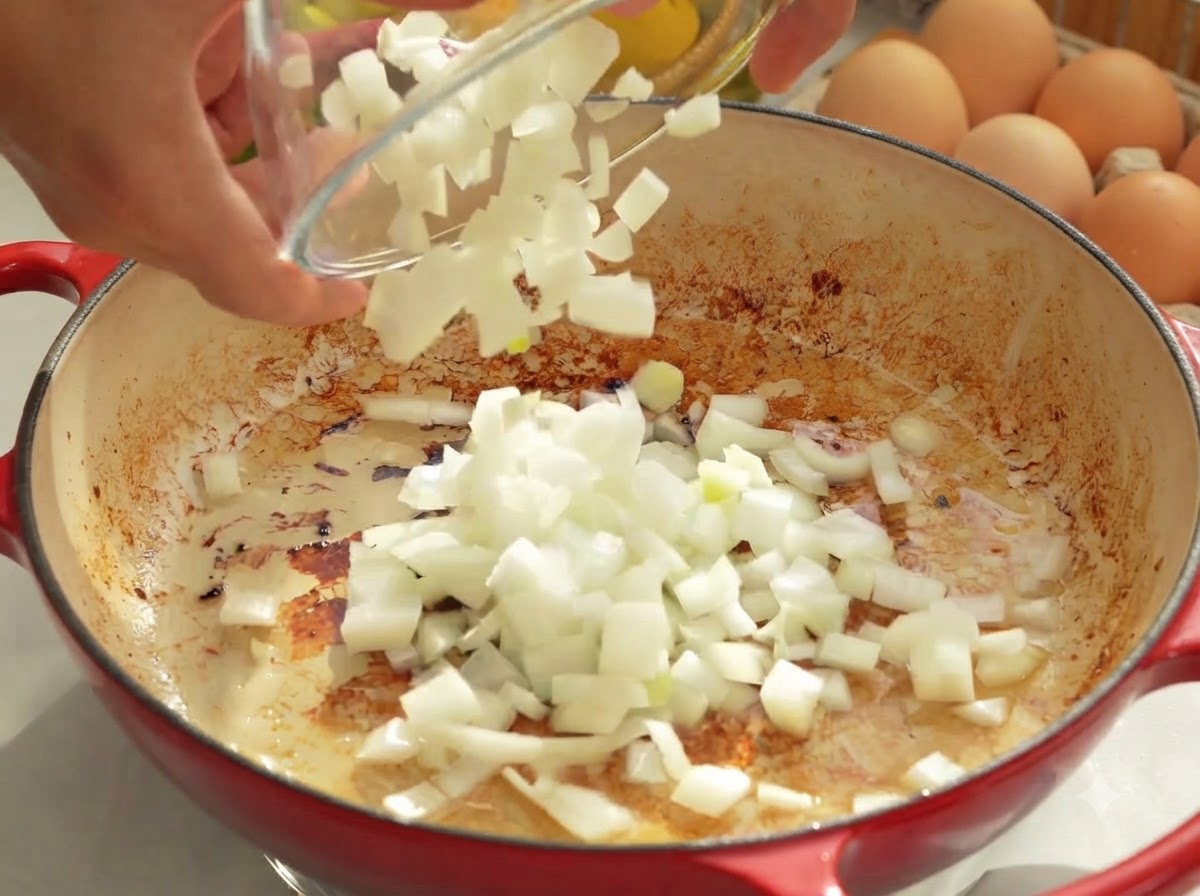 Chopped onions being poured from a glass bowl into a hot pan with rendered fat.