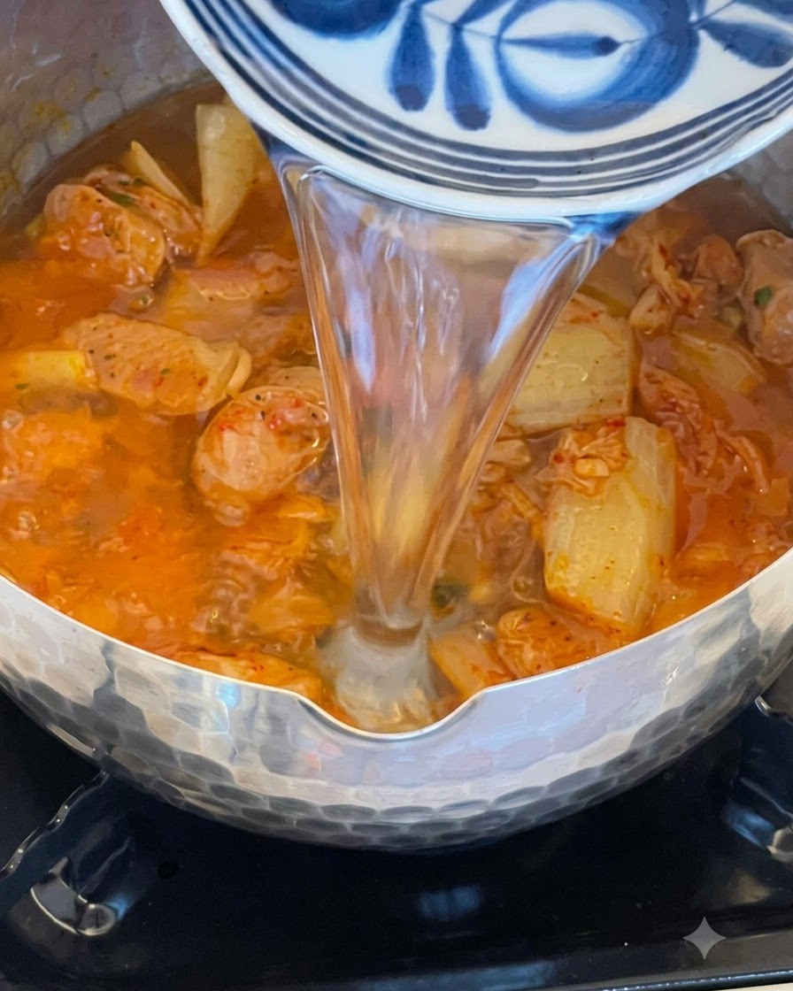 Water pouring from a decorative blue and white bowl into a pot filled with bubbling red kimchi and sausage broth.