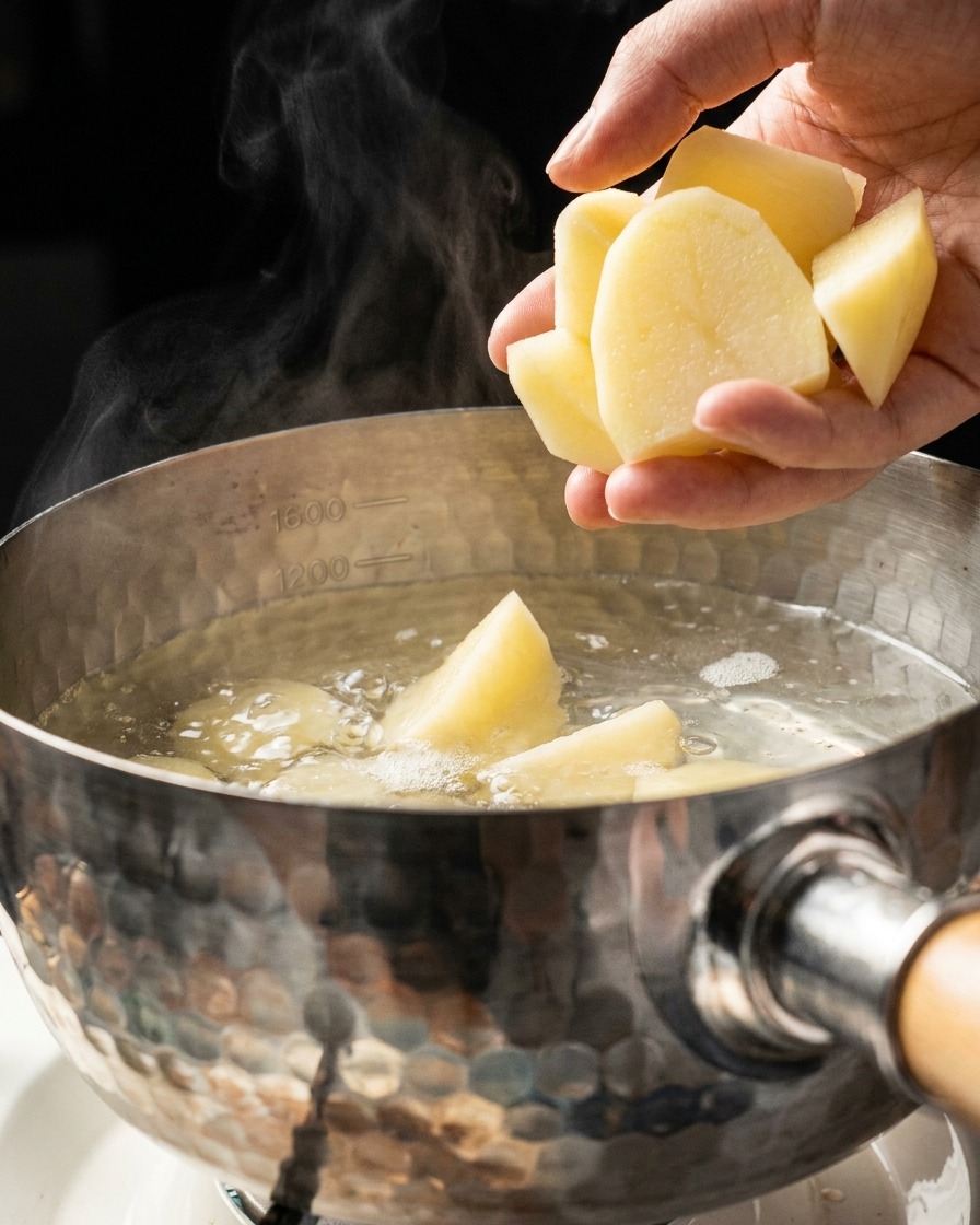 A hand dropping cut potato chunks into a metal pot filled with boiling water and rising steam.