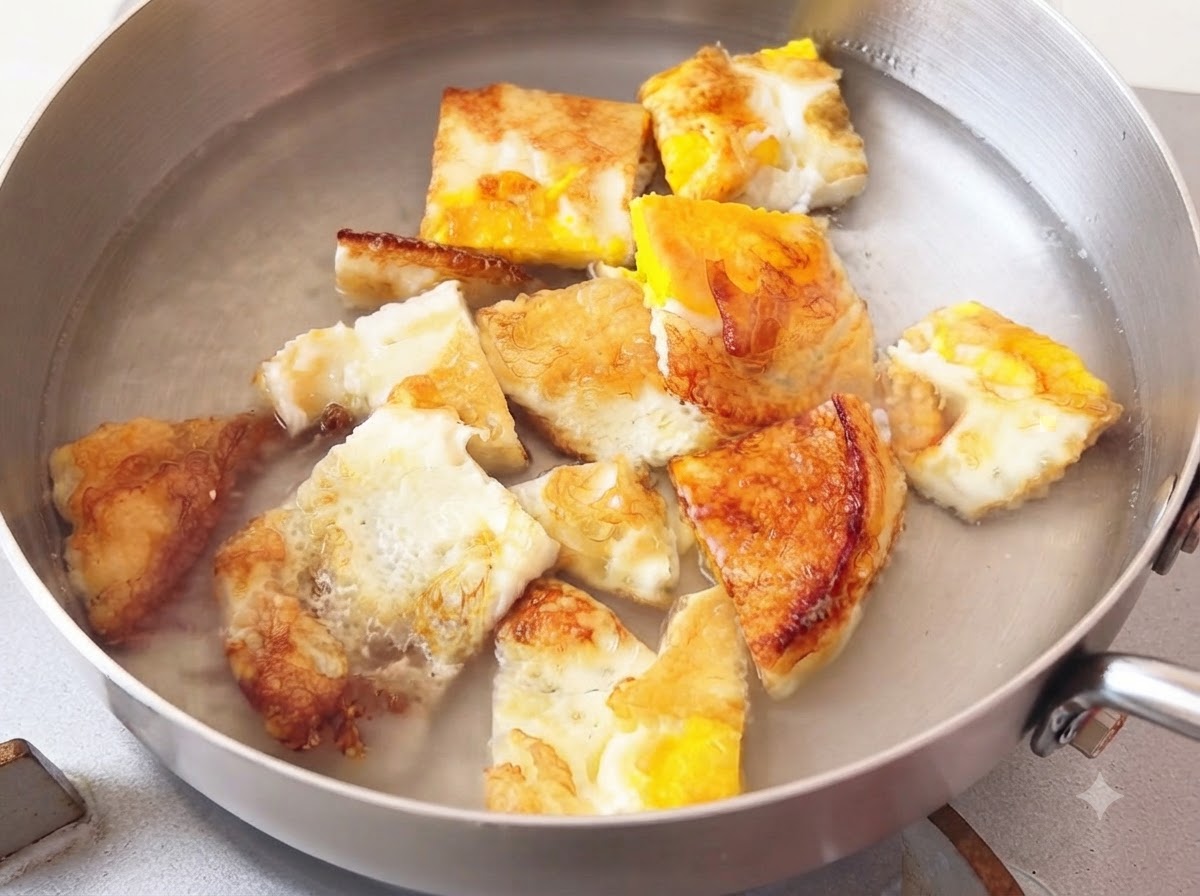 Hot water being poured into a metal cooking pot containing fried egg pieces.