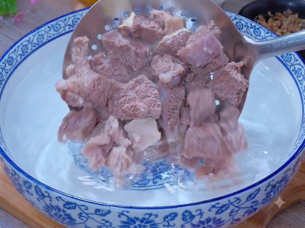 Blanched beef pieces being cleaned inside a bowl of warm water using a slotted spoon.