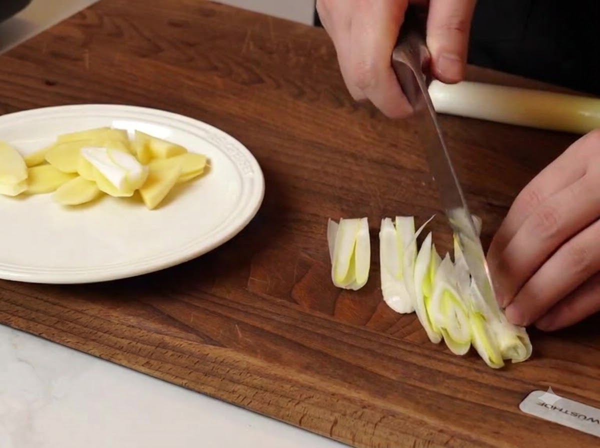 A chef slicing green onions and ginger pieces on a wooden cutting board next to a white plate.