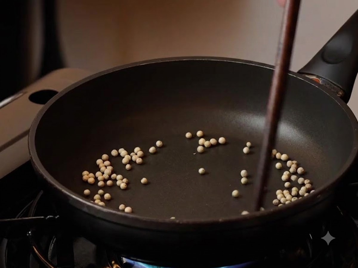 White peppercorns being toasted in a black skillet with chopsticks stirring them over low heat.