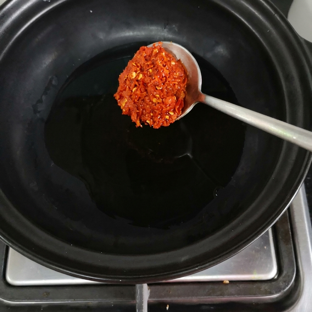 A spoonful of red chili paste being dropped into a black clay pot on the burner.