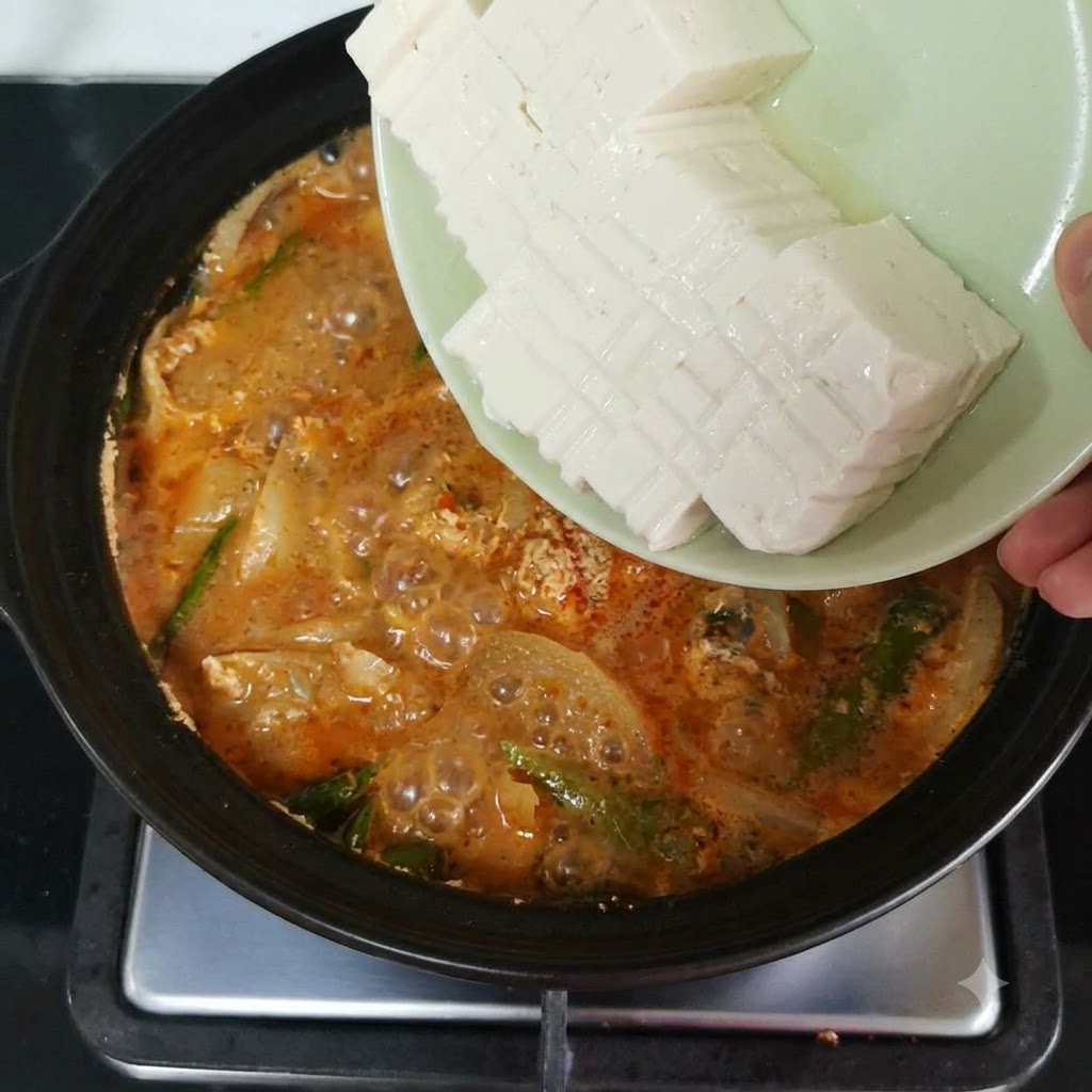 Sliced soft tofu being tipped from a plate into a simmering pot filled with orange-red broth and vegetables.