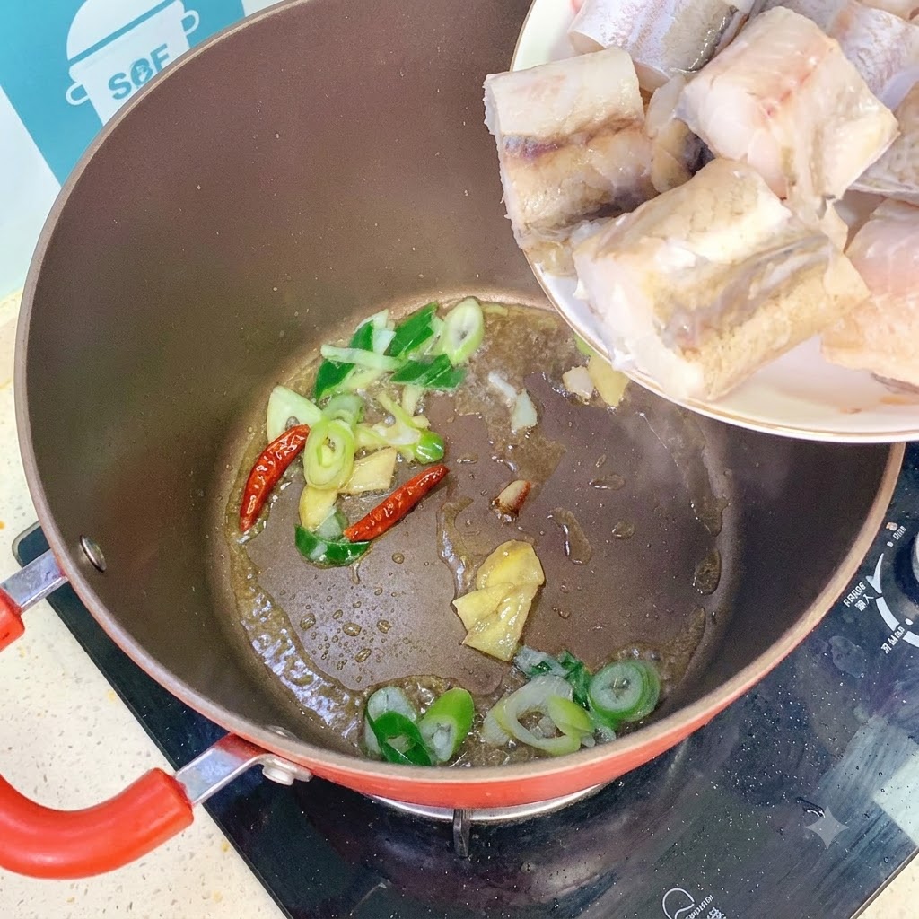 Pollack fish pieces being added from a white plate into a pot containing sautéed scallions, ginger, and dried chilies.