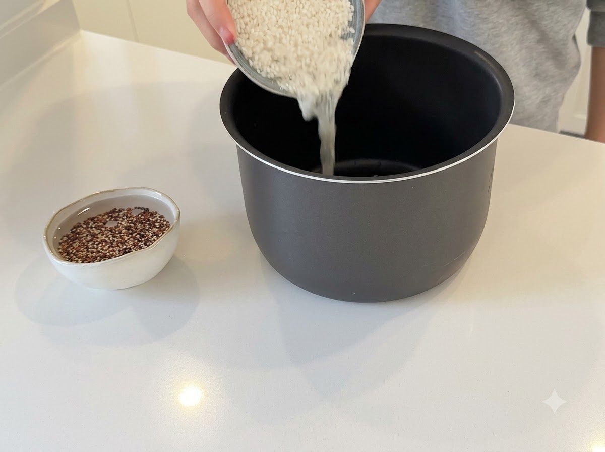 Hands pouring white rice into a dark rice cooker pot with a small bowl of quinoa soaking nearby.