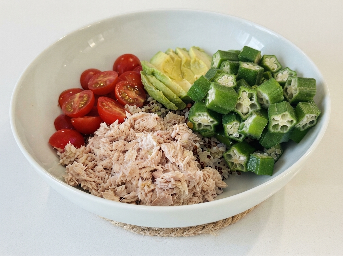 A white bowl filled with a base of rice, topped with sections of tuna, cherry tomatoes, sliced avocado, and okra.
