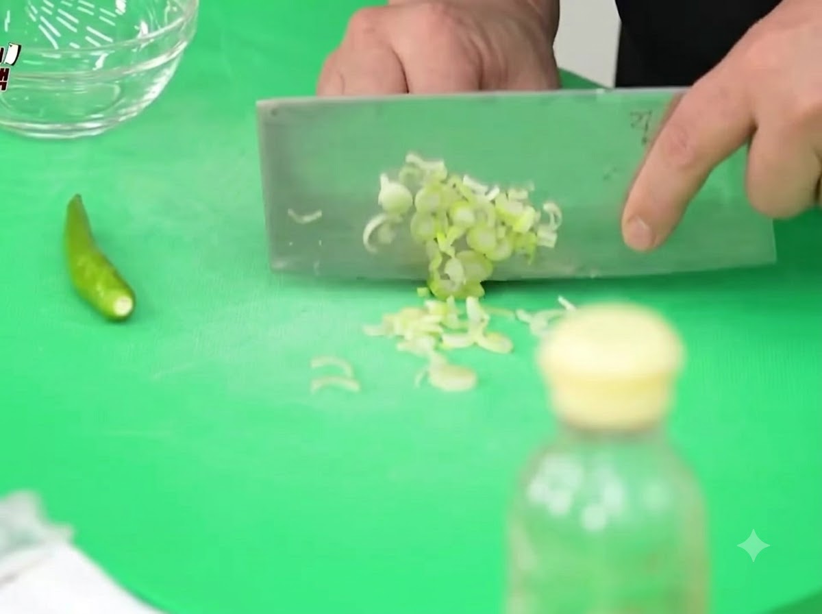 Green onions, Cheongyang chili peppers, and onions being finely chopped on a green cutting board with a cleaver.