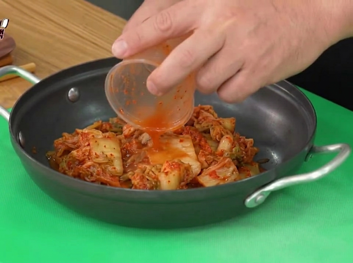 Sour kimchi being added from a plastic cup into a black cooking pot.