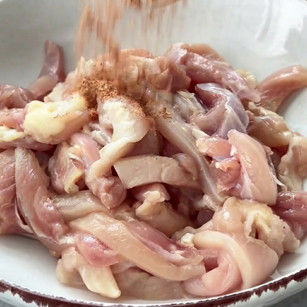 Spices being showered onto a bowl full of raw chicken strips ready to be mixed.