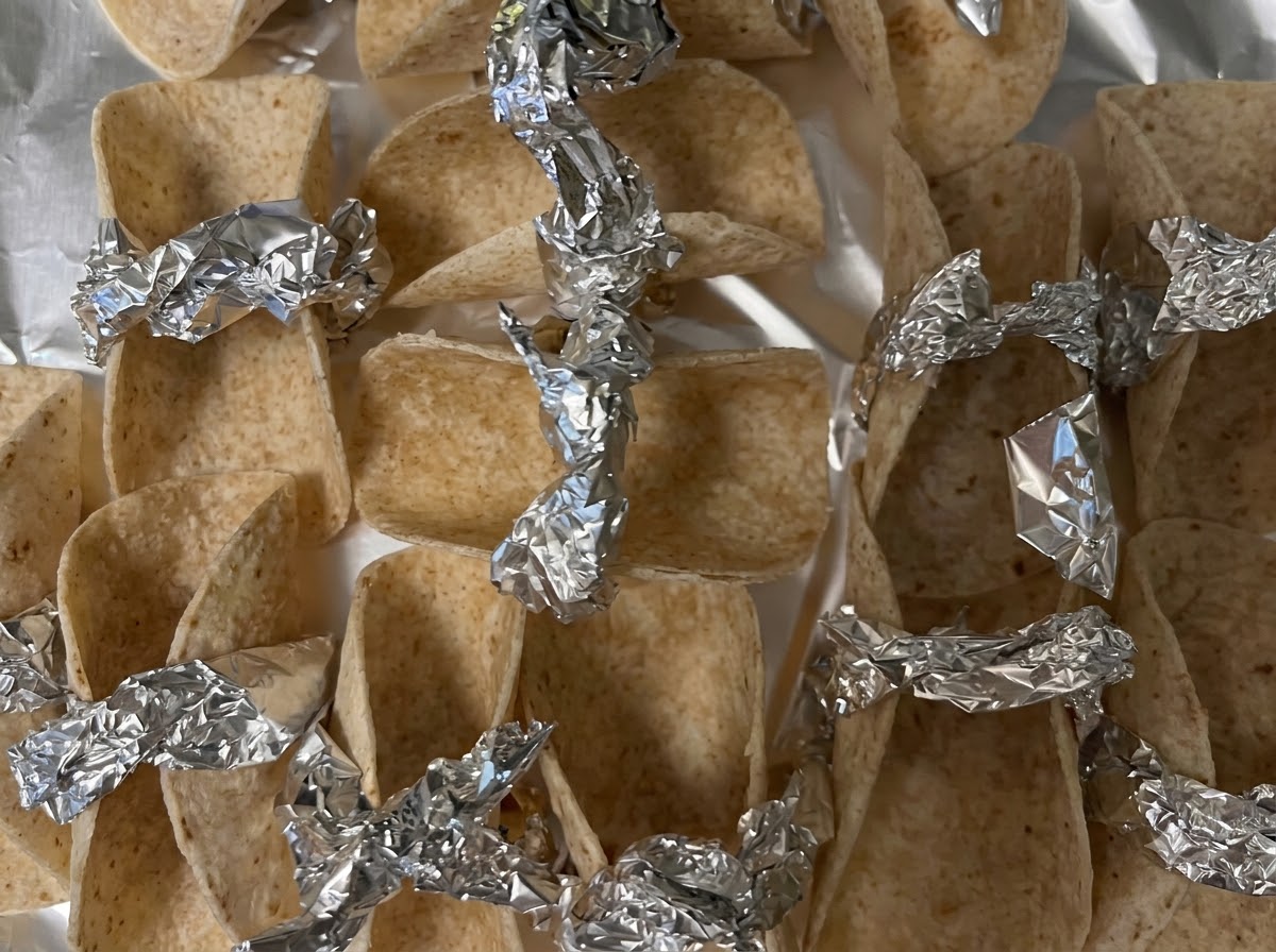 Small tortilla wrappers propped up and secured with foil pieces on a baking sheet ready for the oven.