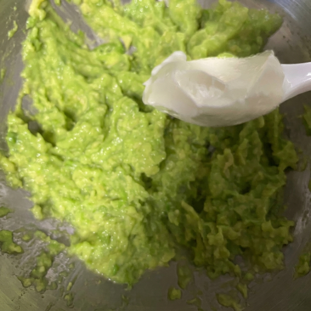 A white spoon scooping thick Greek yogurt and adding it into a bowl of freshly mashed avocado.