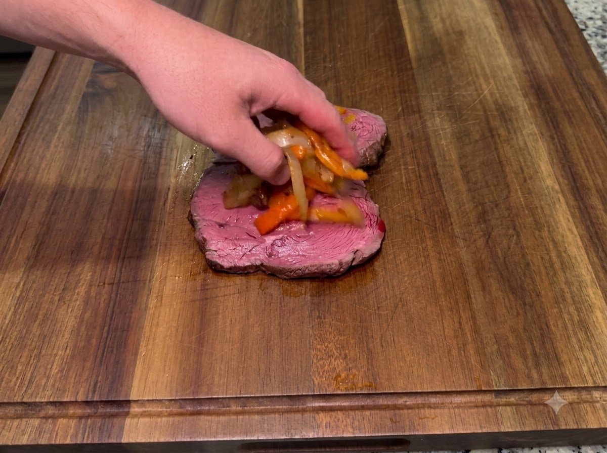 Sautéed onions and smoked spicy peppers being placed on top of a bottom steak piece.