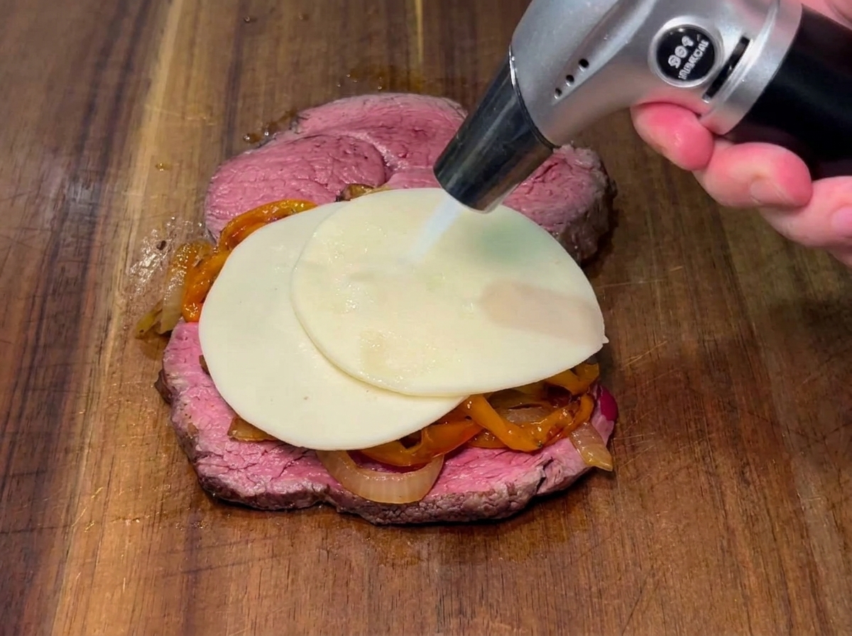 A slice of cheese being melted with a butane torch over peppers and onions on a steak.