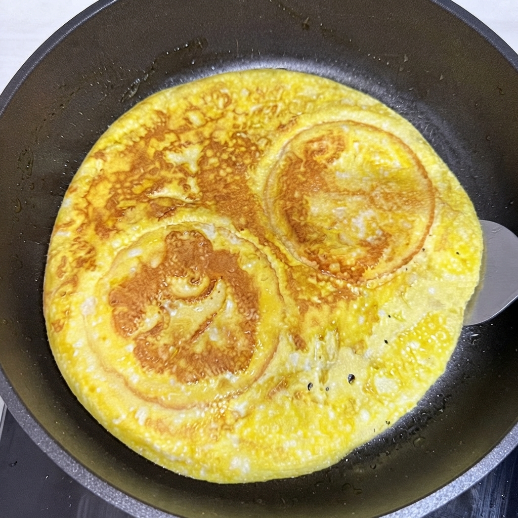 A spatula flipping a round cooked egg and burger bun layer inside a frying pan.