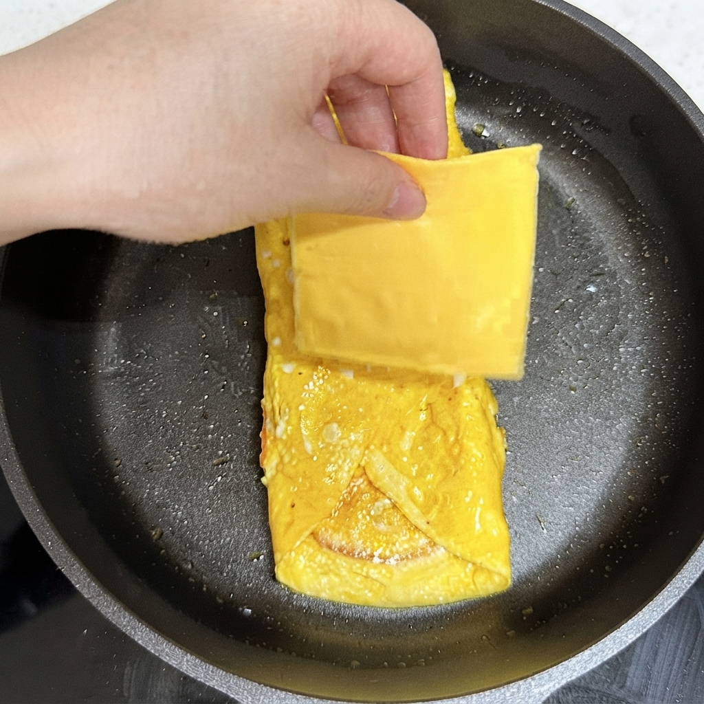 A hand placing a square slice of yellow cheese onto the cooked egg package in the frying pan.