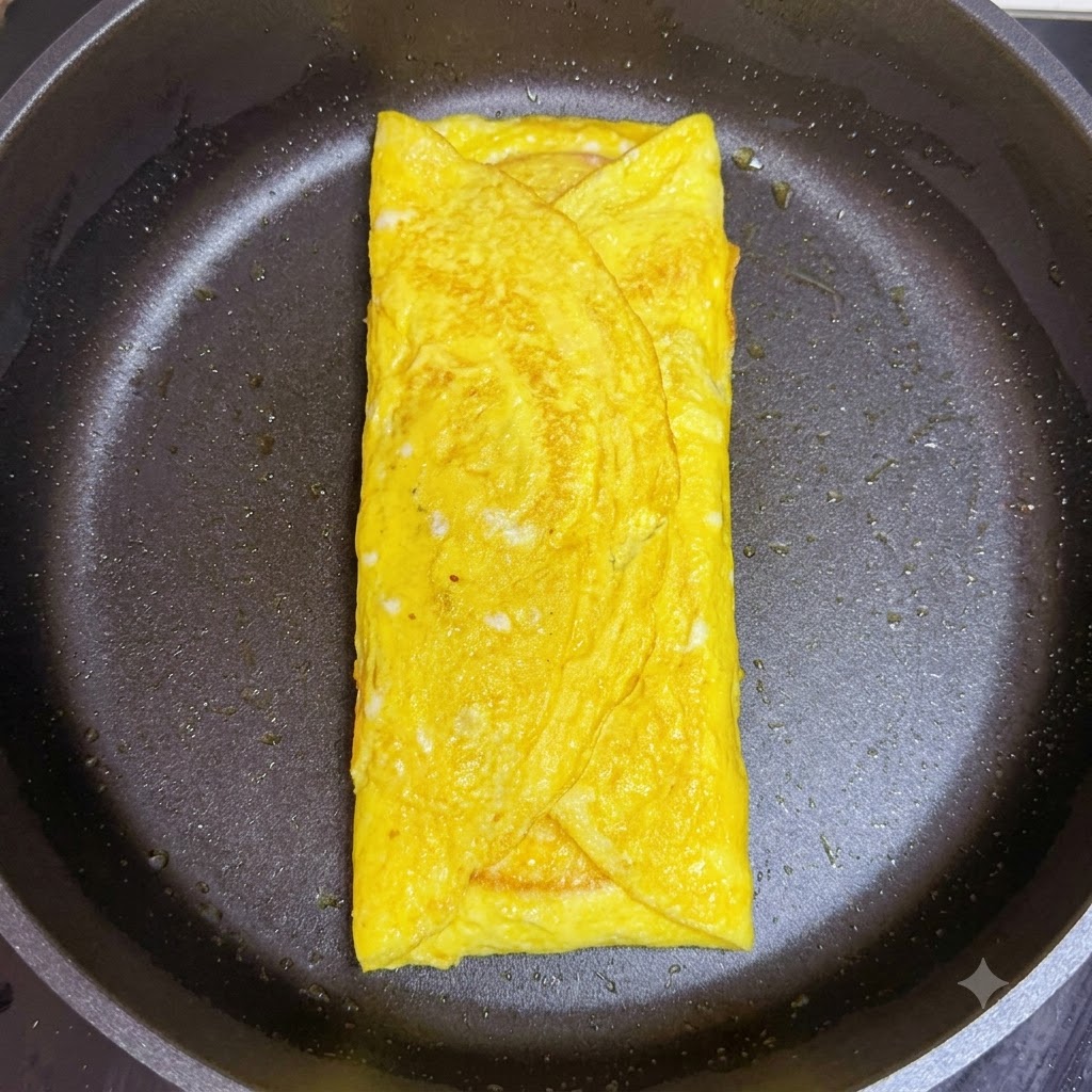 A folded slice of pink ham being layered over the cheese inside the cooked egg wrap in the pan.
