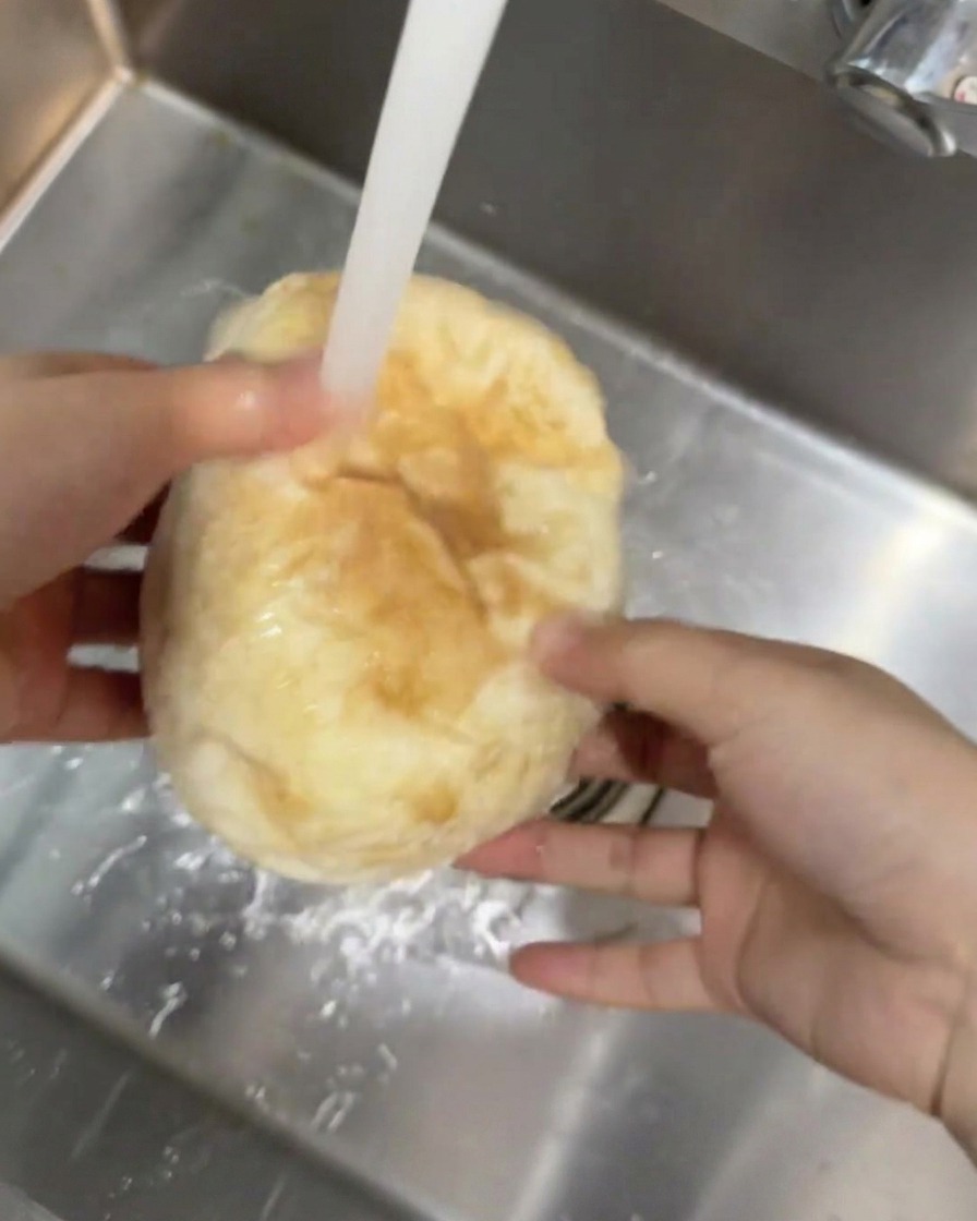 A round ciabatta bread being held under a stream of running tap water in a kitchen sink.
